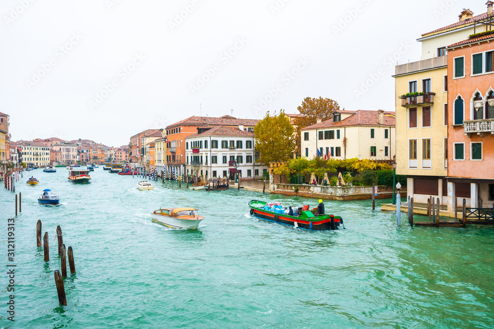 Boats, yachts, vaporettos, water taxis sailing down the Grand Canal waterway between wooden and striped mooring poles and colorful Venetian architecture buildings. November in Venice city, Italy.