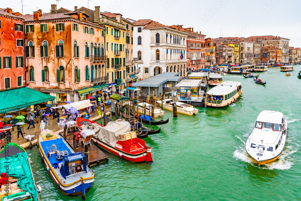 Water taxis, vaporettos, boats, gondolas sailing and docked on Grand Canal along wooden mooring poles and colorful Venetian architecture buildings. People/ tourists in Venice city, Italy on rainy day.