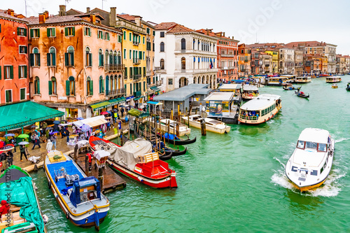 Water taxis, vaporettos, boats, gondolas sailing and docked on Grand Canal along wooden mooring poles and colorful Venetian architecture buildings. People/ tourists in Venice city, Italy on rainy day.