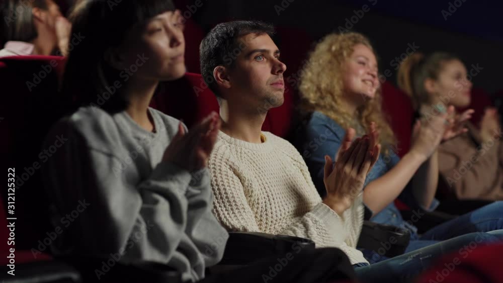 Side view medium shot of satisfied audience applauding after watching movie in cinema, focus on Caucasian young man