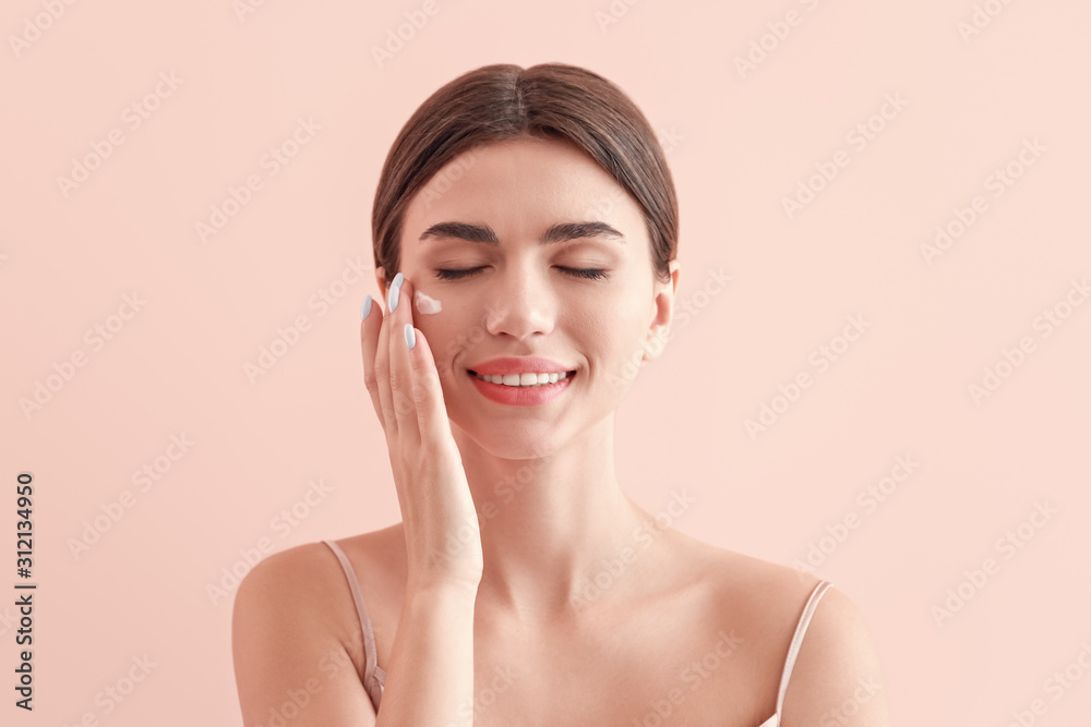 Beautiful young woman applying cream on her face against color background Beautiful young woman applying cream on her face against color background