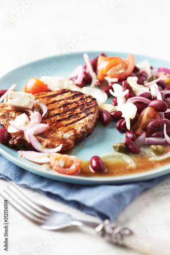 Grilled pork steak with Kidney bean, cherry tomatoes, onion and capers on bright wooden background