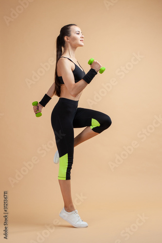A young brunette girl raises her knees high, using extra weights, a full-length photo, against a peach background.