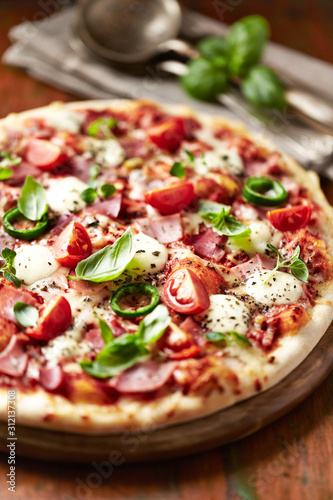 Pizza with ham, cherry tomatoes, mozzarella cheese, jalapeno pepper and fresh herbs. Rustic wooden background