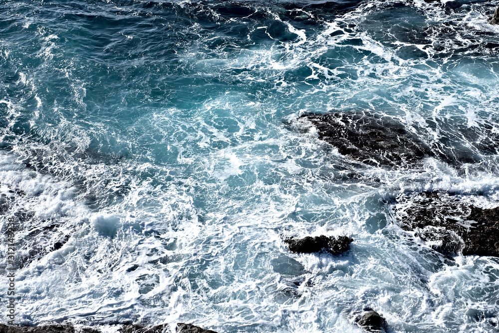 Fototapeta premium Stormy ocean waves, deep blue sea water with white foam and dark dangerous rocks.