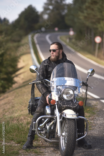 Handsome bearded biker with long hair in leather jacket and sunglasses sitting on cruiser motorcycle on roadside  on blurred background of empty twisty road with white marking on sunny summer day.