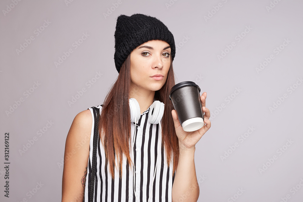 Obraz premium Thoughtful teenage female in knitted hat, headphones on neck making sip from disposable cup with hot coffee, looking seriously at camera, isolated on grey studio blank wall, copy space for advertising