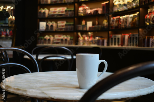 A white coffee mug on a white marble cafe shop table. In the background shelves of candy boxes.