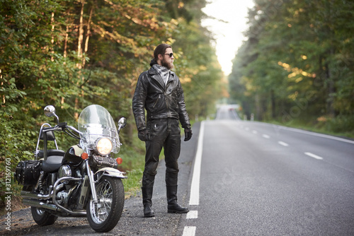 Young bearded tall athletic motorcyclist in black leather outfit and dark sunglasses standing at shiny modern powerful motorbike  on blurred background of hilly asphalt road and green trees.