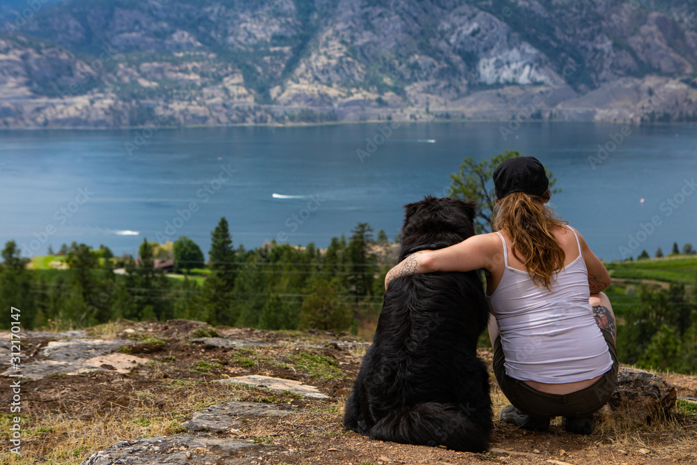 pretty girl over the rocks sits and contemplates the horizon with her beloved dog with a view of the lake of the okanagan valley