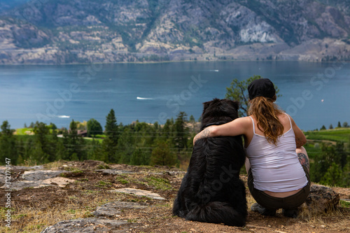 pretty girl over the rocks sits and contemplates the horizon with her beloved dog with a view of the lake of the okanagan valley