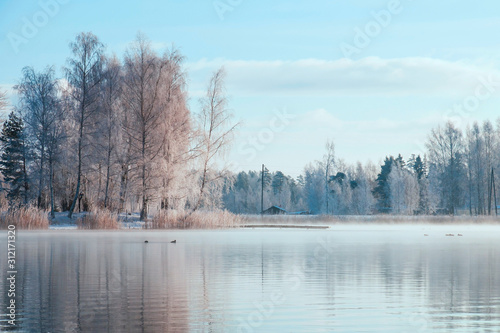 Winter forest on the river at sunset. Colorful landscape with snowy trees, frozen river with reflection in water. Seasonal. Snow covered trees, lake, sun and blue sky.