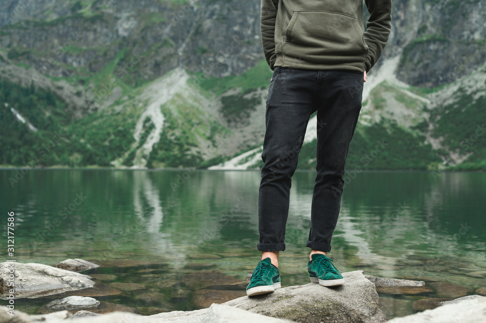 Hiker legs standing on rocks on lake in mountains. A man in sneakers and a green sweater stands on the shores of lake Morskie Oko. Background. Hiker man legs on a hike. Hiker legs standing on rocks on lake in mountains. A man in sneakers and a green sweater stands on the shores of lake Morskie Oko. Background. Hiker man legs on a hike.