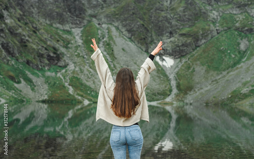 Fototapeta Naklejka Na Ścianę i Meble -  Joyful girl in sweater stands with arms raised on background of lake and mountain, back view. Hipster girl hiking in mountains, posing at camera with raised hands. Woman feels freedom in the mountains