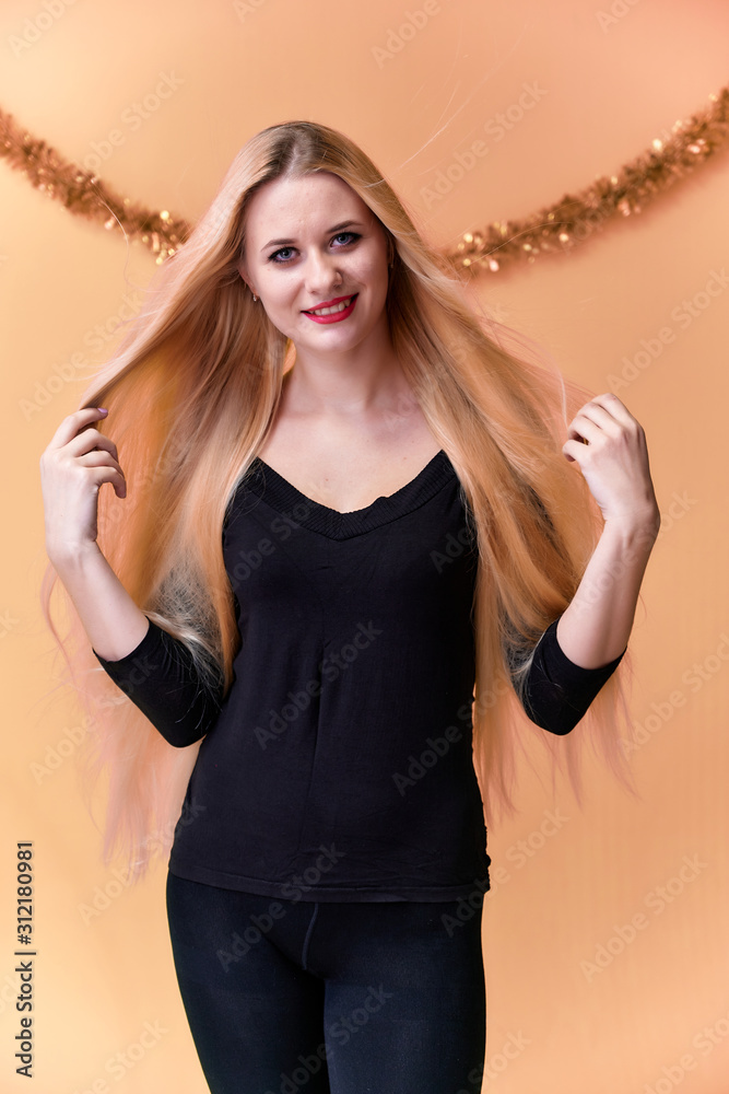 Fototapeta premium Portrait of a cute girl in a black T-shirt with long beautiful hair and great makeup. Concept of a young blonde woman with New Year's decor. Smiling, showing emotions on a pink background.