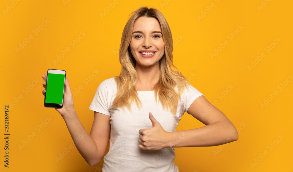 The right choice. Close-up photo of a lovely girl in a white t-shirt, who is looking in the camera with a broad smile and holding a phone in her right hand, while showing thumbs up with her left hand. The right choice. Close-up photo of a lovely girl in a white t-shirt, who is looking in the camera with a broad smile and holding a phone in her right hand, while showing thumbs up with her left hand.