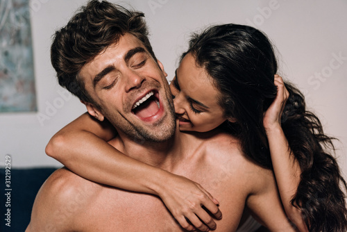 happy woman biting neck of cheerful boyfriend in bedroom