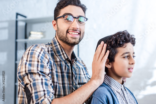Smiling Jewish father wearing hat on son in apartment