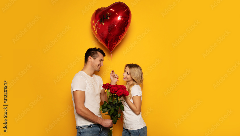 Happy Valentine’s Day. Close-up photo of a lovely couple in casual clothes, who are celebrating Valentine’s Day and holding a bouquet of red roses and a big heart-shaped balloon in their hands. - obrazy, fototapety, plakaty 