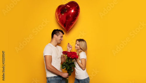 Happy Valentine’s Day. Close-up photo of a lovely couple in casual clothes, who are celebrating Valentine’s Day and holding a bouquet of red roses and a big heart-shaped balloon in their hands.