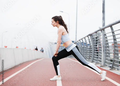 Young sports woman in fitness suit crouches, lunges, plays sports against the backdrop of urban cityscape and white sky.