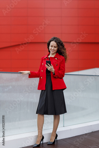Vertical Full length portrait of a elegant business young woman in black dress and red jacket, with mobile phone on hands. Bussines content.