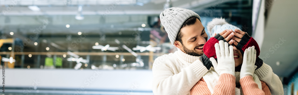 panoramic shot of handsome cheerful man closing eyes to woman to make a surprise on skating rink panoramic shot of handsome cheerful man closing eyes to woman to make a surprise on skating rink
