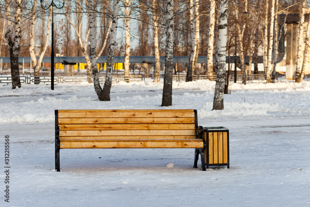 A yellow color painted wooden bench with white beautiful snow and a basket in a park in winter in a sunny day. Russia Bashkortostan