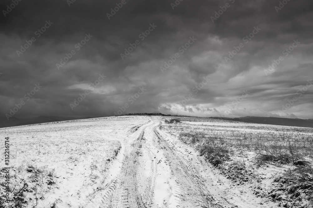 Fototapeta premium Dirt road leading to the top of the hill at wintertime, dramatic storm clouds in black and white.