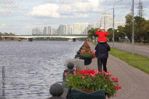 An elderly man with his granddaughter on his shoulders walks along the Nagatinskaya embankment on a summer day against background of Moscow river and metro bridge photo