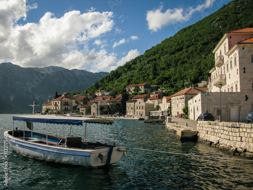 view of the city of Perast in the Bay of Kotor with a boat in the foreground, mountains, blue sky and white clouds