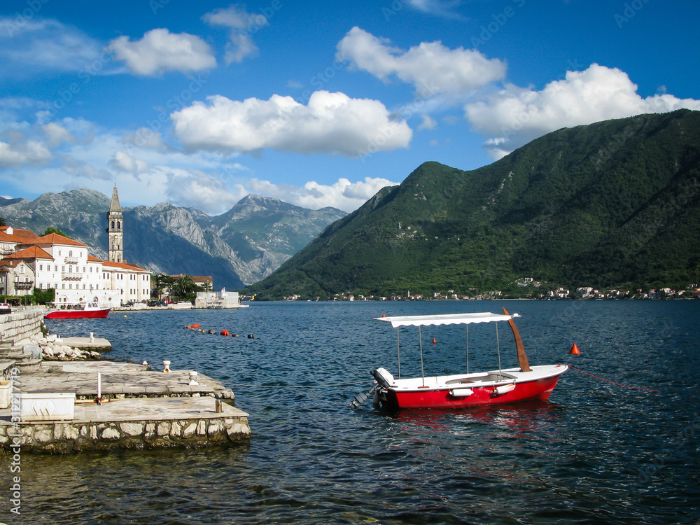 view of the city of Perast in the Bay of Kotor with a boat in the foreground, mountains, blue sky and white clouds