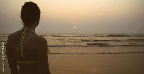 Anonymous woman near sea during sunset. Back view of unrecognizable female in swimwear standing on beach near waving sea in evening on resort