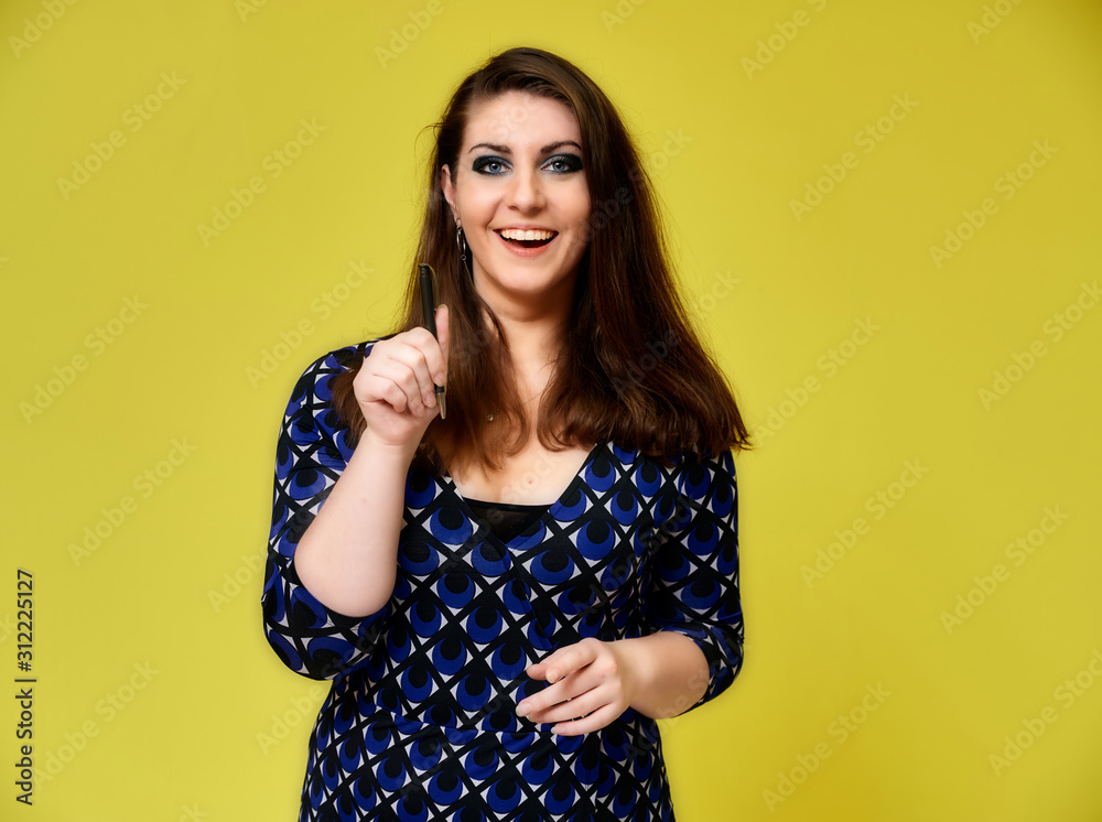 Portrait of a pretty brunette woman with a beautiful hairstyle and with excellent makeup in a dark blue blouse on a yellow background. He stands with a smile and shows with his hands. Portrait of a pretty brunette woman with a beautiful hairstyle and with excellent makeup in a dark blue blouse on a yellow background. He stands with a smile and shows with his hands.