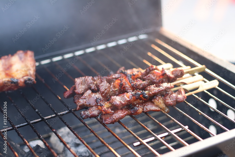 Assorted delicious grilled meat with beef stick and beef steak on black stove with smoke. grills some kind of marinated meat and vegetable on gas grill during summer party time. Assorted delicious grilled meat with beef stick and beef steak on black stove with smoke. grills some kind of marinated meat and vegetable on gas grill during summer party time.
