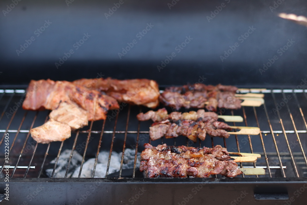 Assorted delicious grilled meat with beef stick and beef steak on black stove with smoke. grills some kind of marinated meat and vegetable on gas grill during summer party time. 