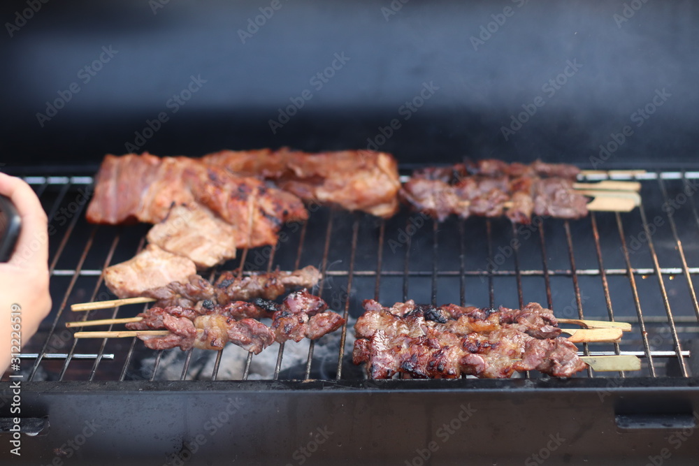 Assorted delicious grilled meat with beef stick and beef steak on black stove with smoke. grills some kind of marinated meat and vegetable on gas grill during summer party time. 