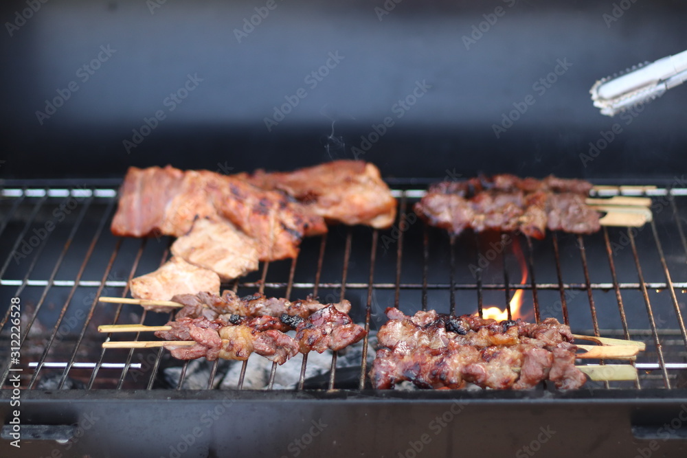 Assorted delicious grilled meat with beef stick and beef steak on black stove with smoke. grills some kind of marinated meat and vegetable on gas grill during summer party time. 