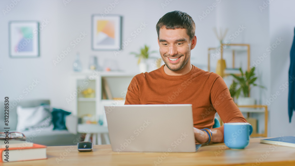 Portrait of Handsome Smiling Man Working on Laptop, Sitting at His Wooden Desk at Home. Man Browsing Through Internet, Working on Notebook from His Living Room. Portrait of Handsome Smiling Man Working on Laptop, Sitting at His Wooden Desk at Home. Man Browsing Through Internet, Working on Notebook from His Living Room.