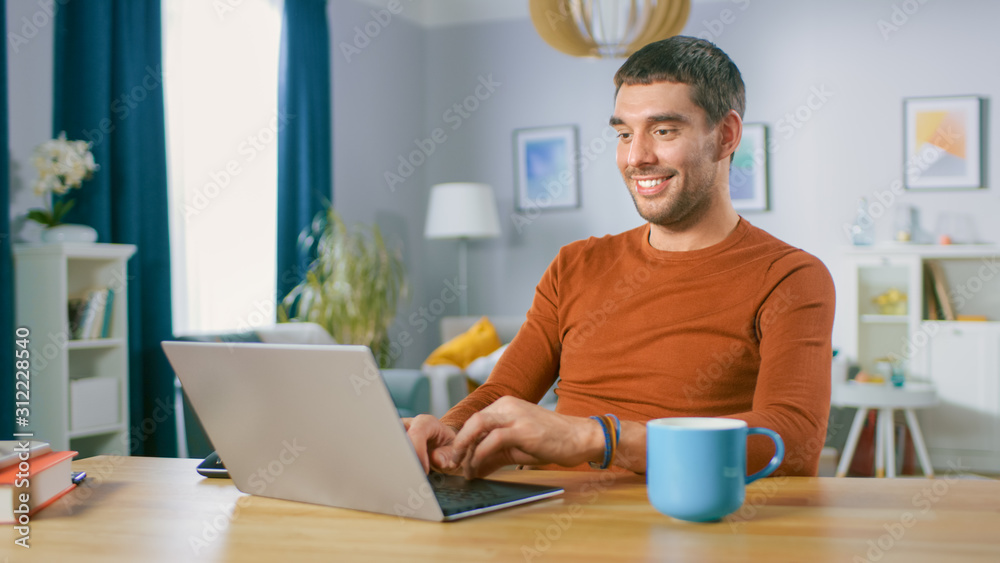 Portrait of Handsome Smiling Man Working on Laptop, Sitting at His Wooden Desk at Home. Man Browsing Through Internet, Working on Notebook from His Living Room. Portrait of Handsome Smiling Man Working on Laptop, Sitting at His Wooden Desk at Home. Man Browsing Through Internet, Working on Notebook from His Living Room.