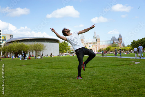 Young man with sunglasses, jeans and white t-shirt, jumping in the grass. A sunny day in Museumplein the museum park in Amsterdam, Holland. People outdoors in spring.