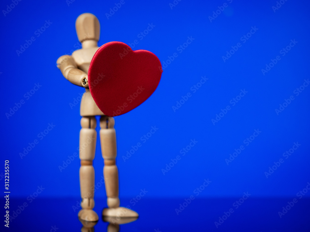 Wooden male figure standing and holding red heart in his hands. Blue background, Copy space. Saint Valentine theme background. Wooden male figure standing and holding red heart in his hands. Blue background, Copy space. Saint Valentine theme background.