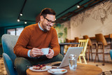 Portrait of a smiling businessman having coffee and slice of cake and looking at laptop.