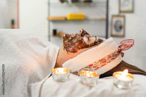 Young woman with coffee face treatment at spa salon cosmetologist mask for peeling on the face of a girl in a cosmetology room cosmetic therapy lying on bed head covered with towel