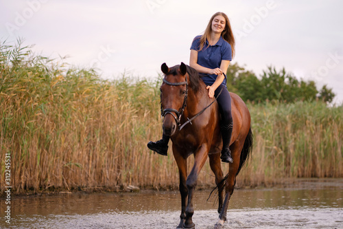 A young girl riding a horse on a shallow lake. A horse runs on water at sunset. Care and walk with the horse. Strength and Beauty © Kate