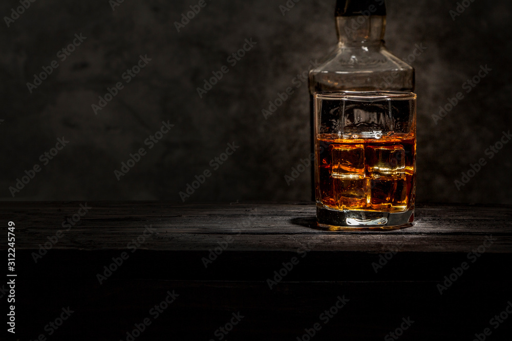 Whiskey with ice or brandy in a glass and a square carafe on an old wooden background. Whiskey with ice in a glass. Whiskey or cognac. Selective focus. Whiskey with ice or brandy in a glass and a square carafe on an old wooden background. Whiskey with ice in a glass. Whiskey or cognac. Selective focus.