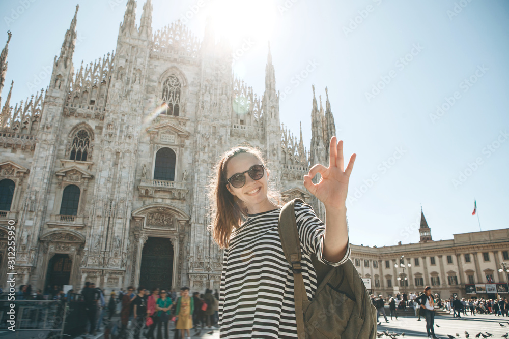 Fototapeta premium Positive girl tourist or student with a backpack on the background of the Duomo Cathedral in Milan in Italy shows a hand sign meaning cool.