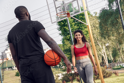 Outdoors Activity. African couple standing on basketball court man back view with ball looking at woman walking towards smiling cheerful