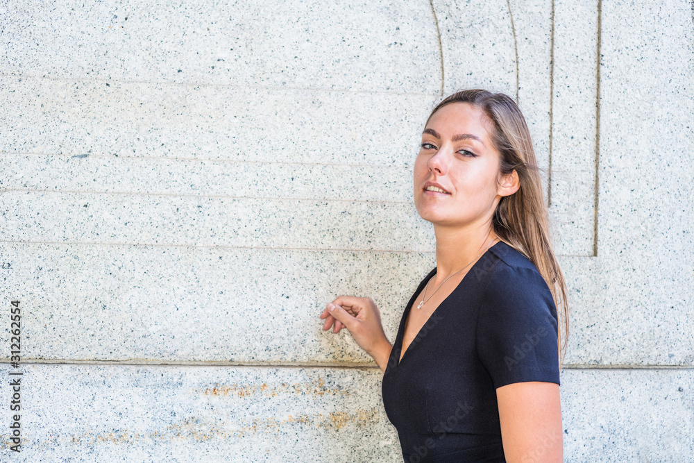 Portrait of Young East European Woman in New York City. Young college female student with long brown hair, wearing black short sleeve, v neck dress, standing by wall on street, looking forward..