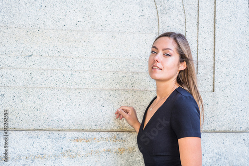 Obraz na płótnie Portrait of Young East European Woman in New York City. Young college female student with long brown hair, wearing black short sleeve, v neck dress, standing by wall on street, looking forward.. w salonie
